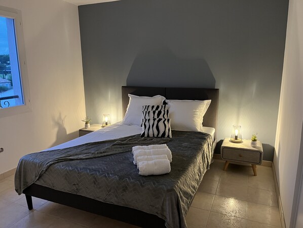 A neatly made bed with a dark patterned bedspread, white pillows, and a decorative pillow with a zebra print. There are two bedside lamps on either side of the bed, and a small potted plant on the nightstand.
