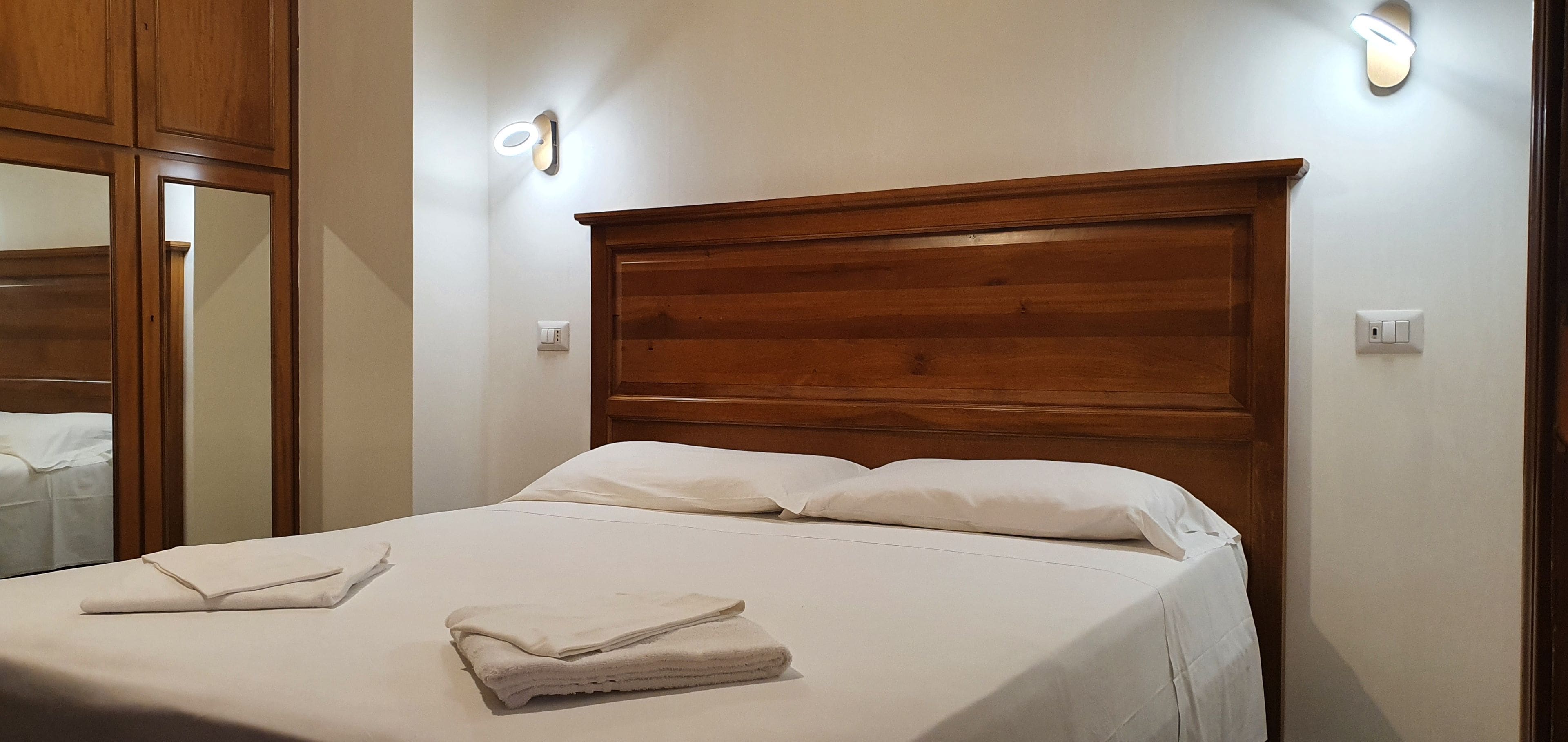 Una cama bien hecha con sábanas blancas y toallas dobladas, cabecera de madera y lámparas fijadas a la pared.