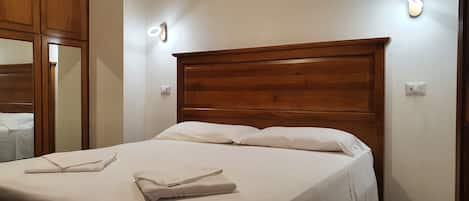 Una cama bien hecha con sábanas blancas y toallas dobladas, cabecera de madera y lámparas fijadas a la pared.