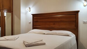Una cama bien hecha con sábanas blancas y toallas dobladas, cabecera de madera y lámparas fijadas a la pared.