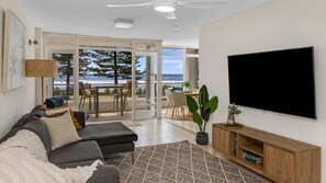 Een woonkamer met een grijze bank, een flatscreen televisie en een houten kast. Er is een groot raam met uitzicht op de oceaan en bomen.