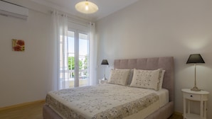 A bedroom with a large bed, two bedside tables with lamps, a window with curtains, and a small wall art piece. - The Bright Suite (Corfu)
