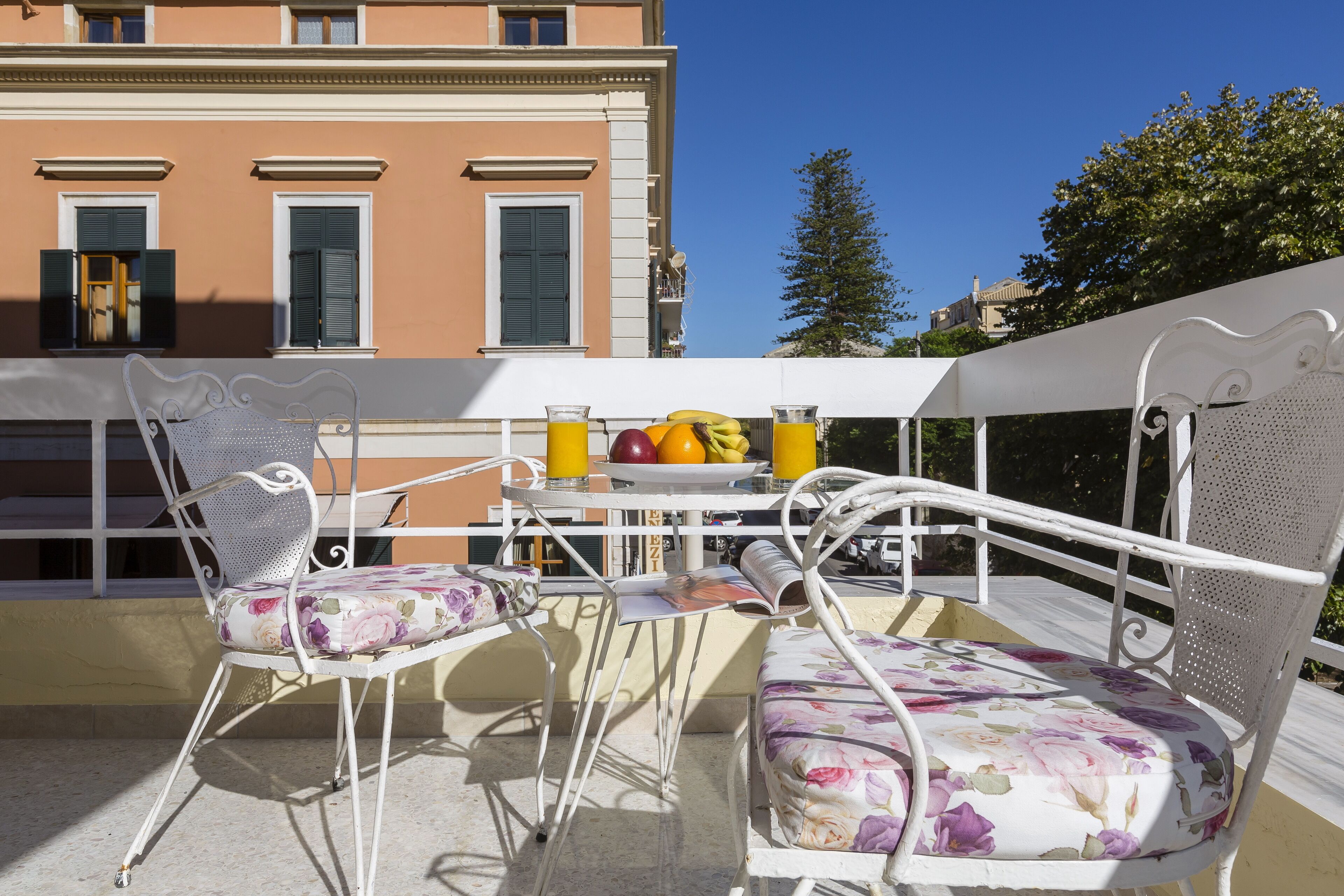 A balcony with white metal chairs and a table, a fruit bowl, and a glass of orange juice.