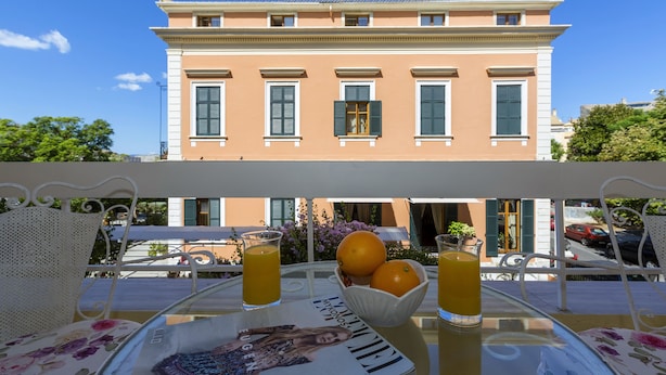 A table with a magazine, a bowl of oranges, and glasses of orange juice, set against a building with a balcony. - The Bright Suite (Corfu)