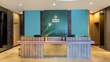 A reception desk at Malya Bhagra Hotel with a modern design, featuring a teal wall with the hotel's logo, a wooden reception counter, and a potted plant.