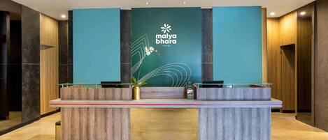A reception desk at Malya Bhagra Hotel with a modern design, featuring a teal wall with the hotel's logo, a wooden reception counter, and a potted plant.