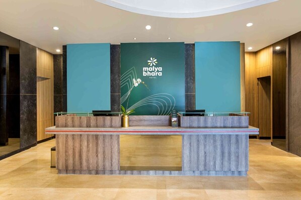 Un mostrador de recepción en el Hotel Malya Bhagra con un diseño moderno, que presenta una pared verde aguamarina con el logotipo del hotel, un mostrador de madera y una planta en maceta.
