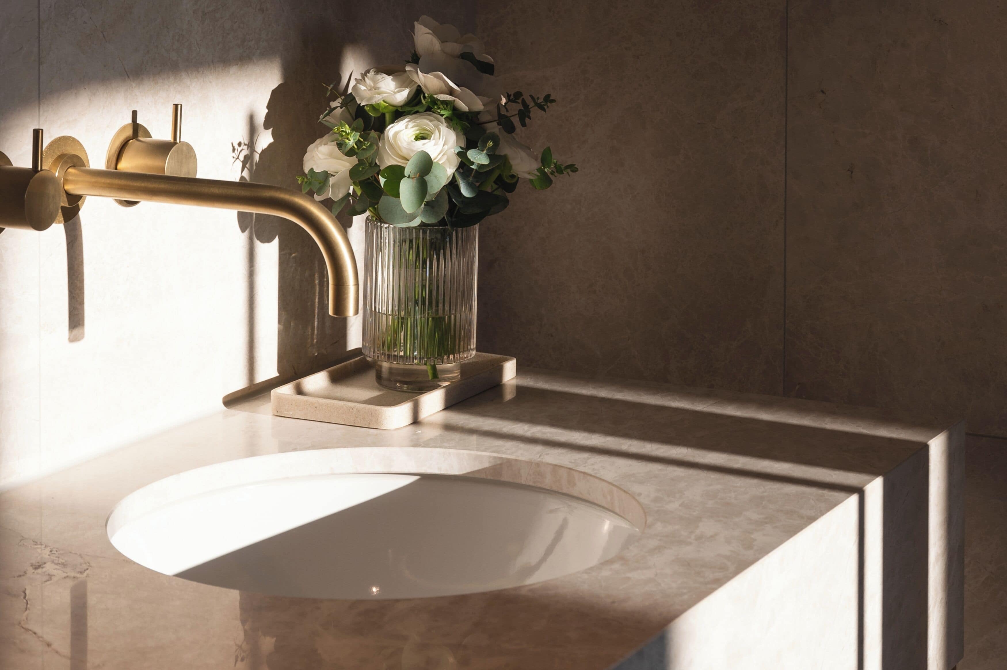 Un baño con grifo dorado, lavamanos blanco y un jarrón de flores blancas sobre una encimera de mármol.
