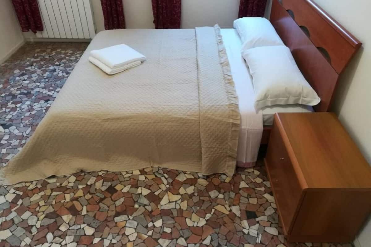 Una cama bien hecha con una colcha y almohadas color beige, una mesita de noche de madera y un piso de mosaicos.