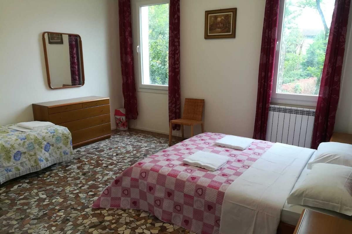Un dormitorio con dos camas, un aparador, una silla, un ventanal con cortinas, un radiador y un cuadro en la pared.