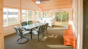 A wooden cabin with a dining table and chairs, a ceiling fan, and large windows offering a view of the outdoors. - Cozy 2 bedroom cottage, lakefront private beach (Township of Branch)