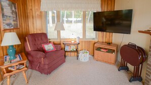 A cozy room with a wooden paneled wall, a red armchair, a television, a small table with a lamp, and a window with a view of trees. - Cozy 2 bedroom cottage, lakefront private beach (Township of Branch)