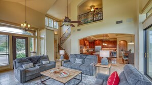 A spacious living area with a high ceiling, a staircase, and a kitchen in the background.