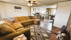 A modern living room with a sectional sofa, a patterned rug, a ceiling fan, and a kitchen in the background.