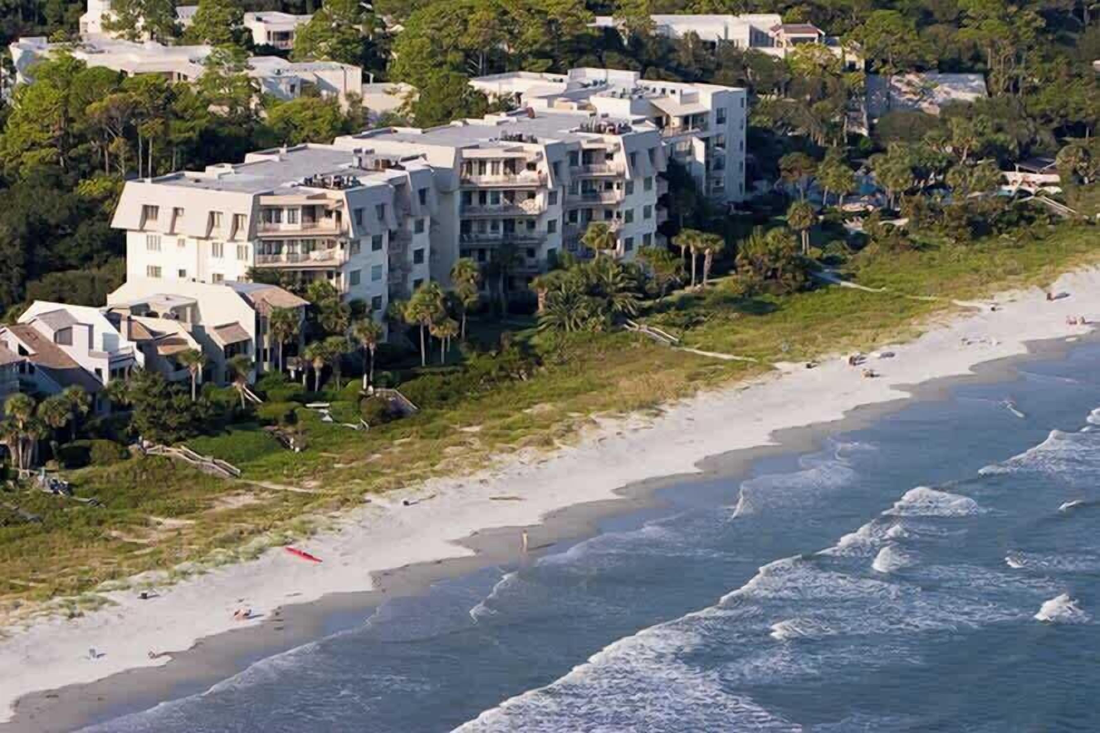 Una zona residencial costera con propiedades frente al mar, incluyendo edificios de varios pisos con balcones, una playa arenosa y olas oceánicas.