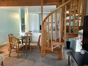 A dining area with a wooden staircase, a kitchen in the background, and a view of the outdoors.