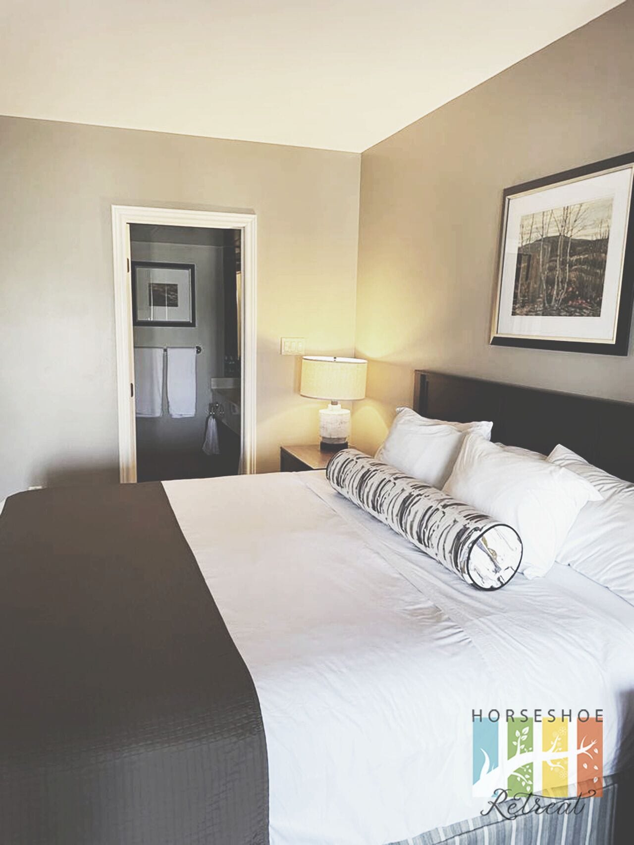 A hotel room with a large bed, a nightstand with a lamp, a framed picture on the wall, and a door leading to a bathroom.