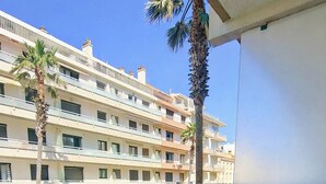 Un balcón con una mesa de plástico blanco, una silla amarilla con cojín estampado, un tapete tejido, un cactus en maceta y un edificio con múltiples balcones al fondo.