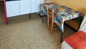 Una cocina compacta con armarios blancos, una pequeña mesa de comedor con mantel colorido y un sofá con un cojín rojo.