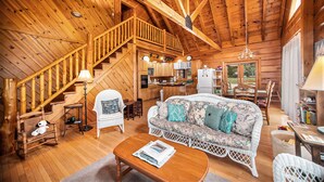 A cozy cabin interior with a wooden staircase, a living room with a sofa and chairs, a dining area, and a kitchen.