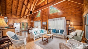 A log cabin interior with a wooden ceiling, a dining area, a living room with wicker furniture, and a kitchen with a refrigerator.