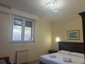 A hotel room with a bed, a window with blinds, a radiator, a nightstand, and a wall-mounted light.