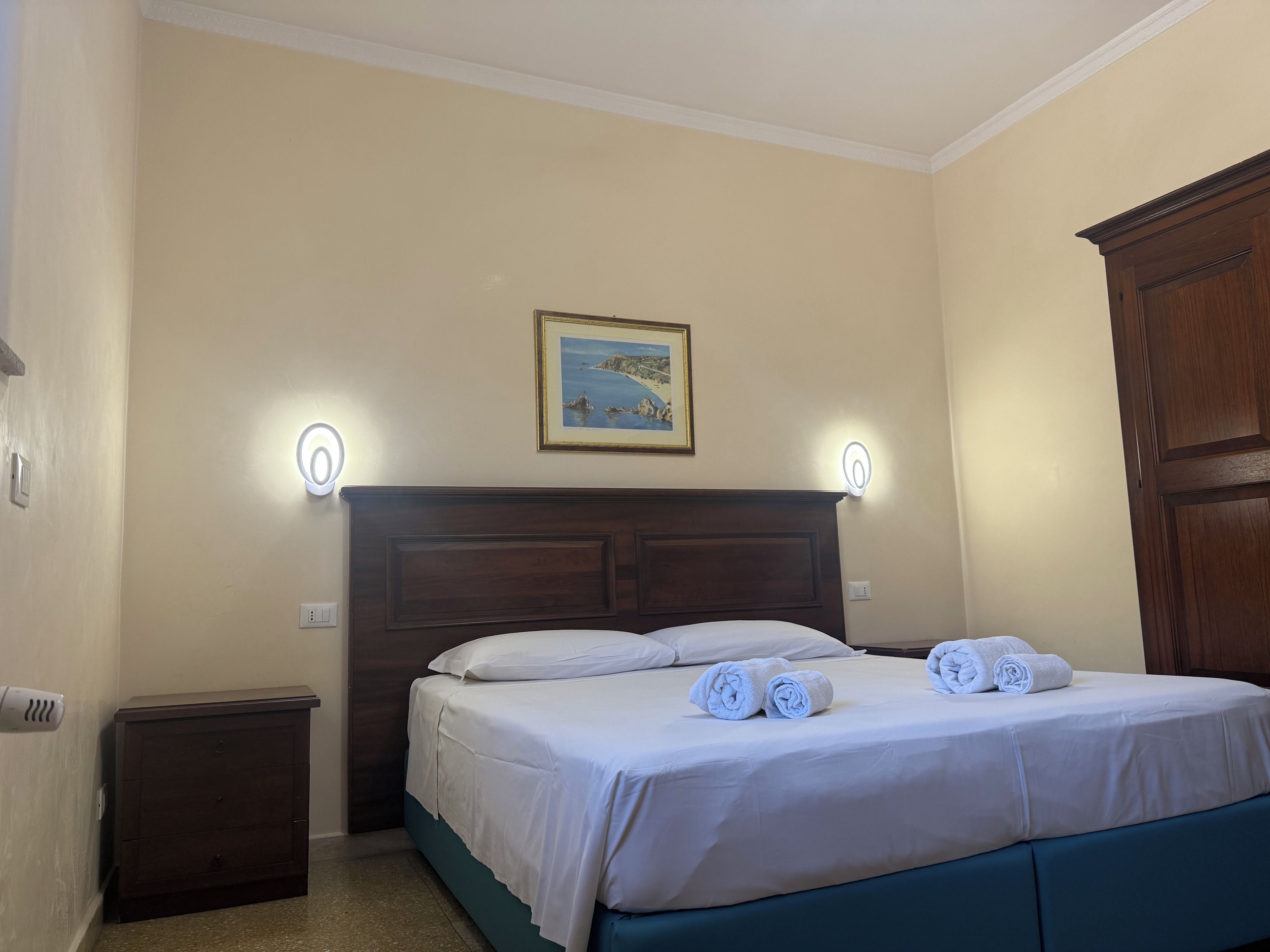 A hotel room with a bed, a nightstand, a wooden wardrobe, and a framed picture on the wall.
