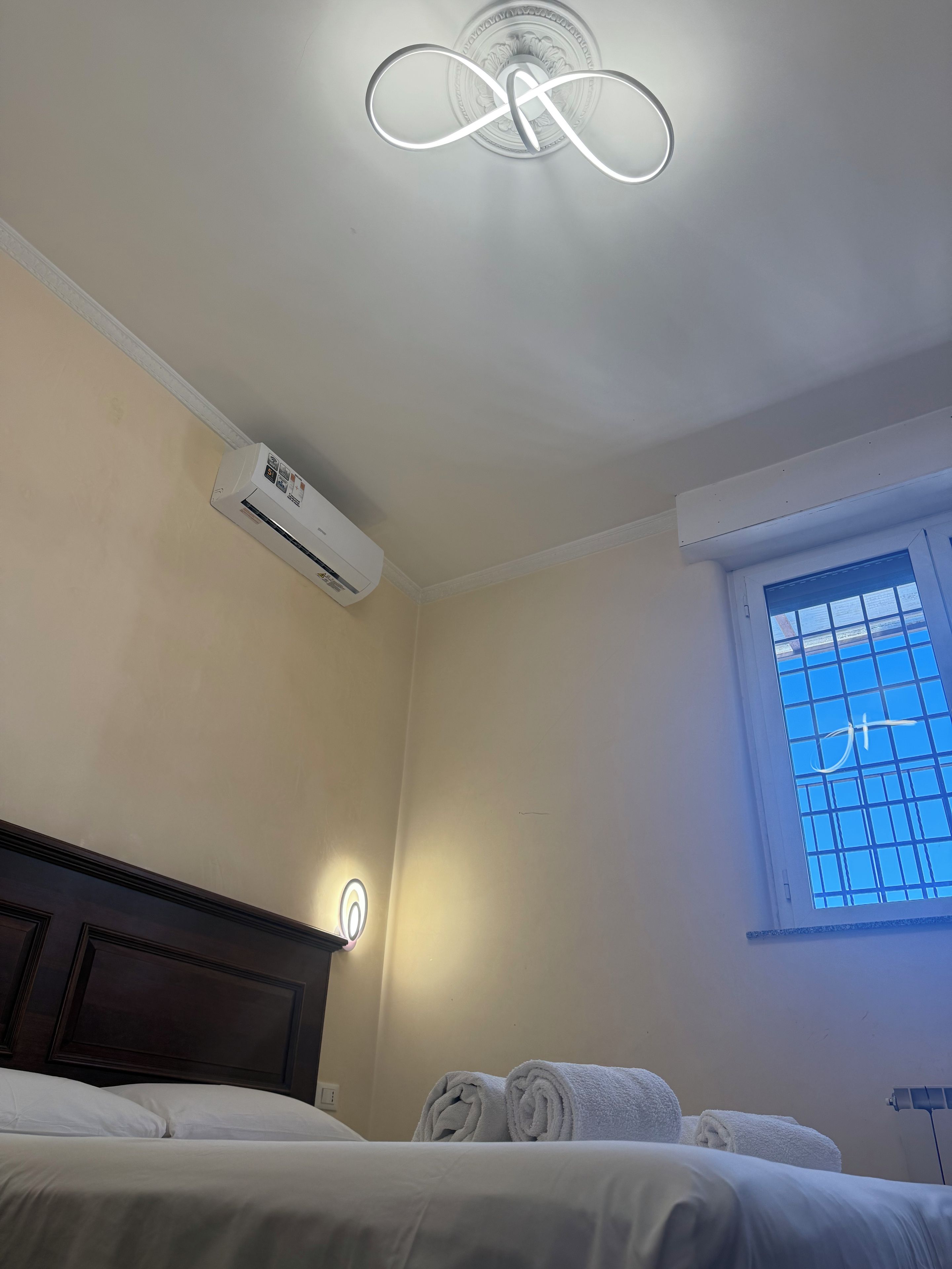 A hotel room with a bed, a wall-mounted air conditioner, a window with a view, and a ceiling light fixture.