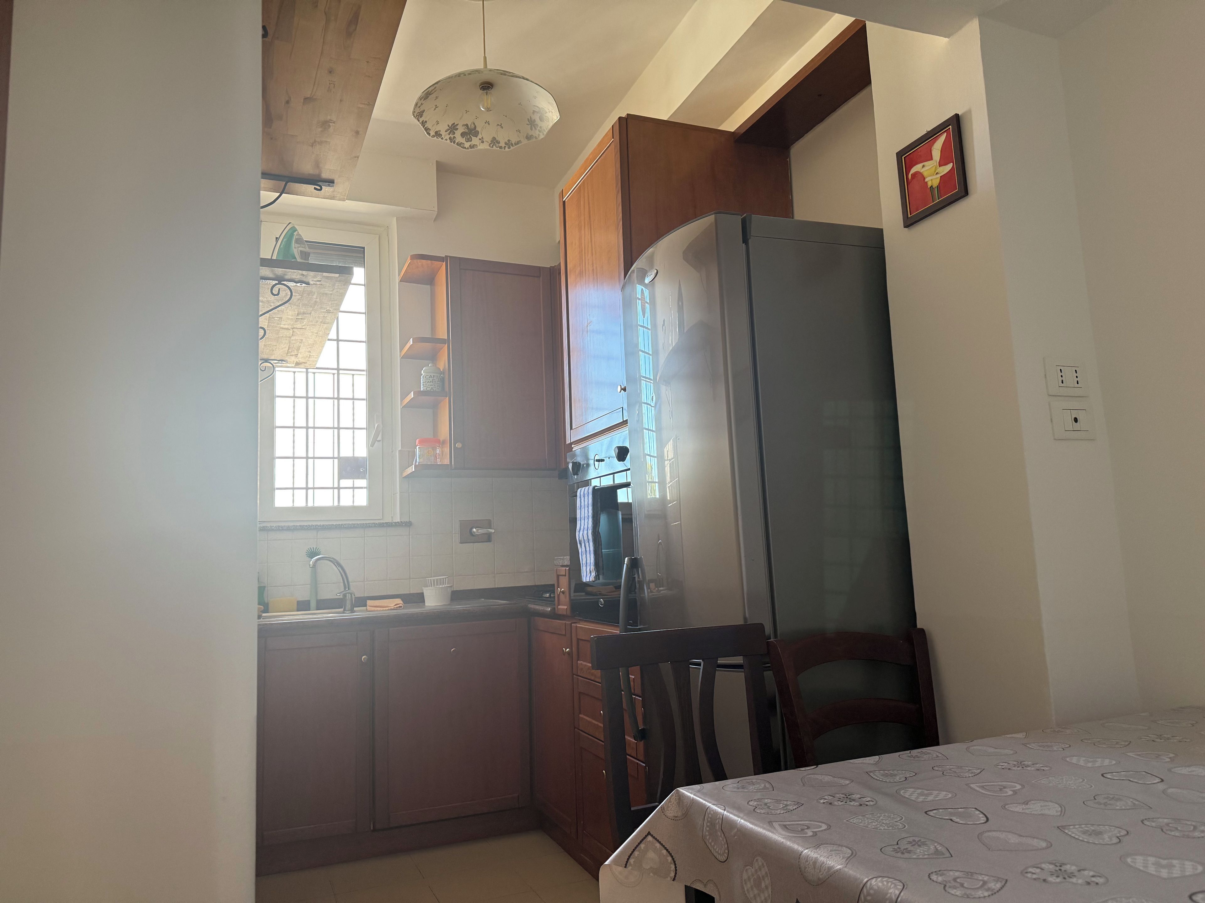 Cocina compacta con muebles de madera, un refrigerador y una mesa de comedor con mantel estampado.