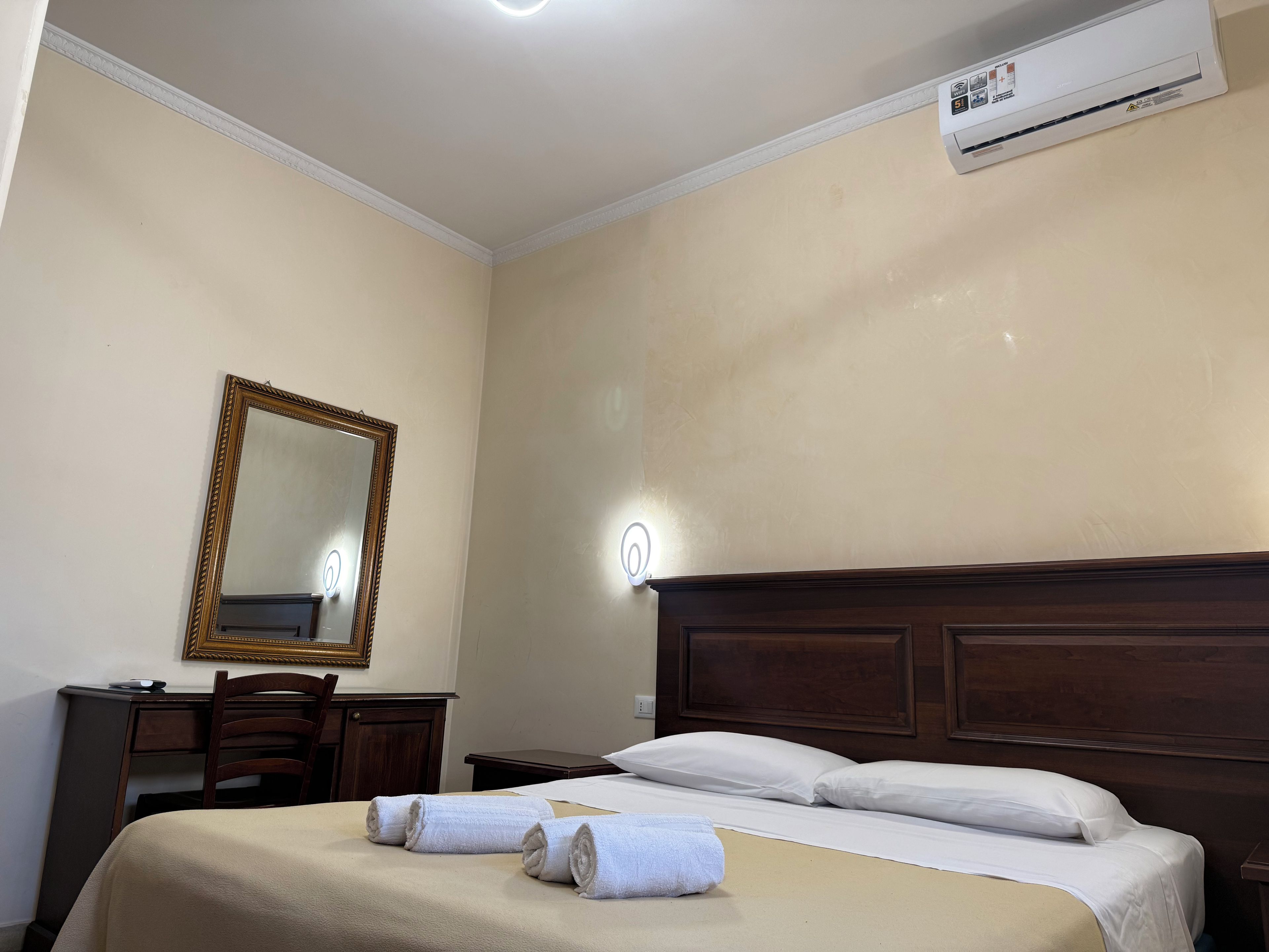 A hotel room with a bed, a desk, a chair, a mirror, and an air conditioning unit.