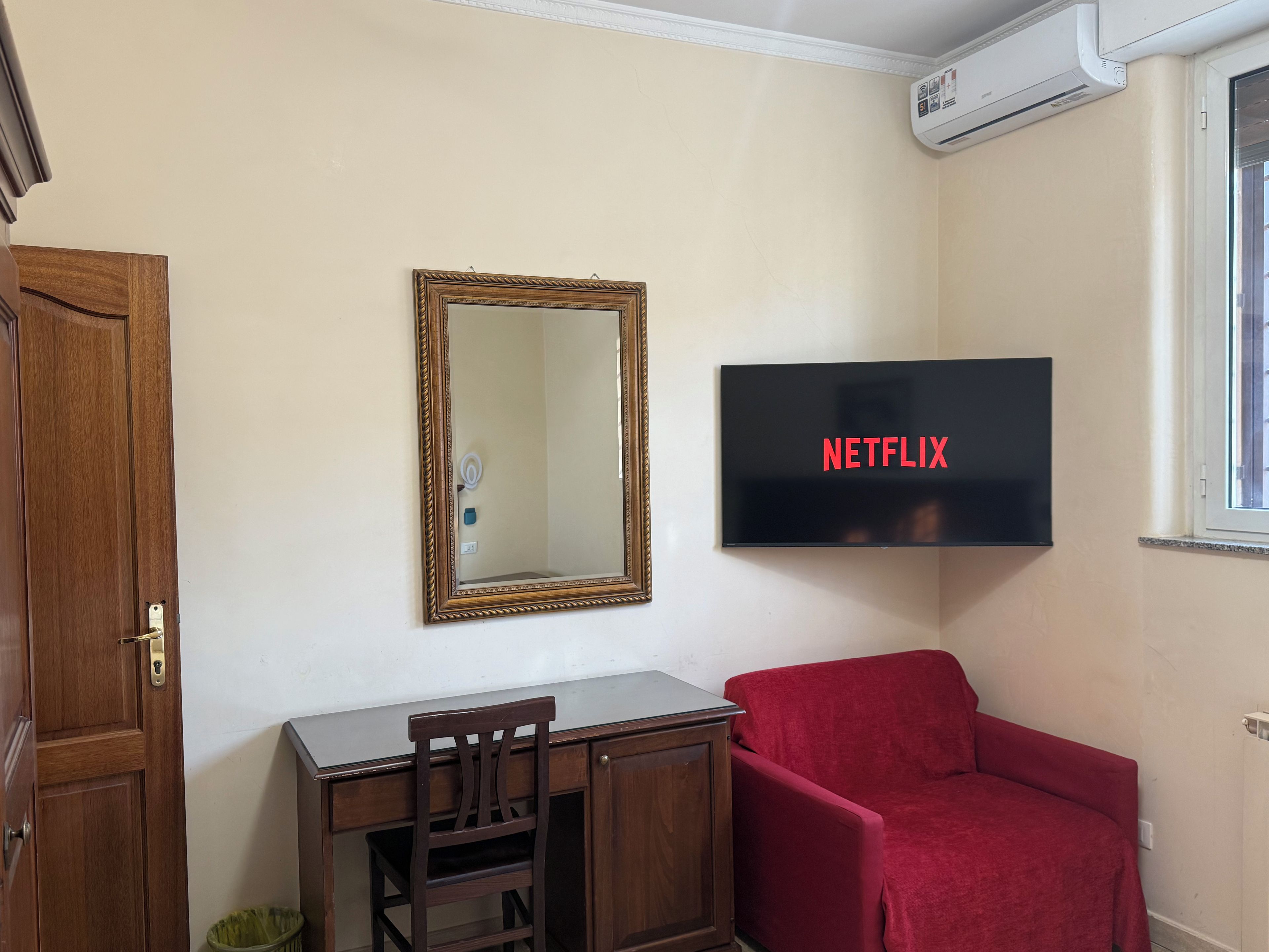 Una habitación con un escritorio de madera, una silla roja, un televisor de pared que muestra 'NETFLIX' y un espejo.