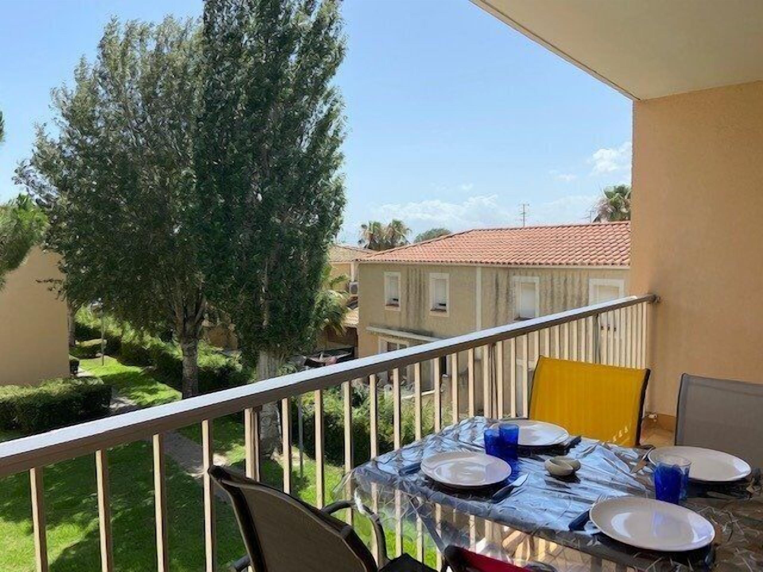 A balcony with a table set for two, overlooking a garden and neighboring buildings.