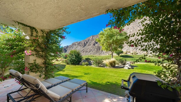 A patio with two lounge chairs, a grill, and a view of a well-maintained garden and mountains.