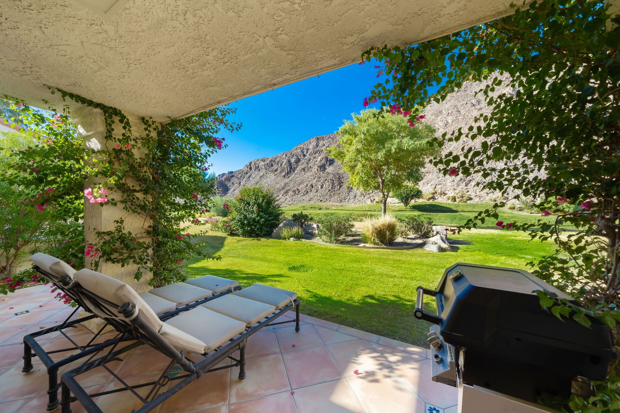 A patio with two lounge chairs, a grill, and a view of a well-maintained garden and mountains.