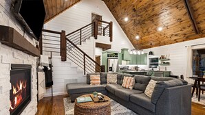 Ein gemütliches Wohnzimmer mit Kamin, einem grauen Ecksofa, einem gewebten Hocker und einer Treppe, die zu einer oberen Ebene führt.