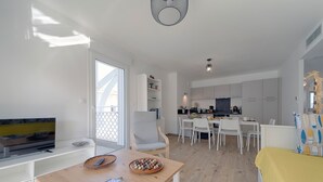 A modern, well-lit living room with a TV, dining table, and chairs. A kitchen is visible in the background with cabinets and a dining area. - Charming apartment ideally located - Swimming pool - Air conditioning (Mimizan)