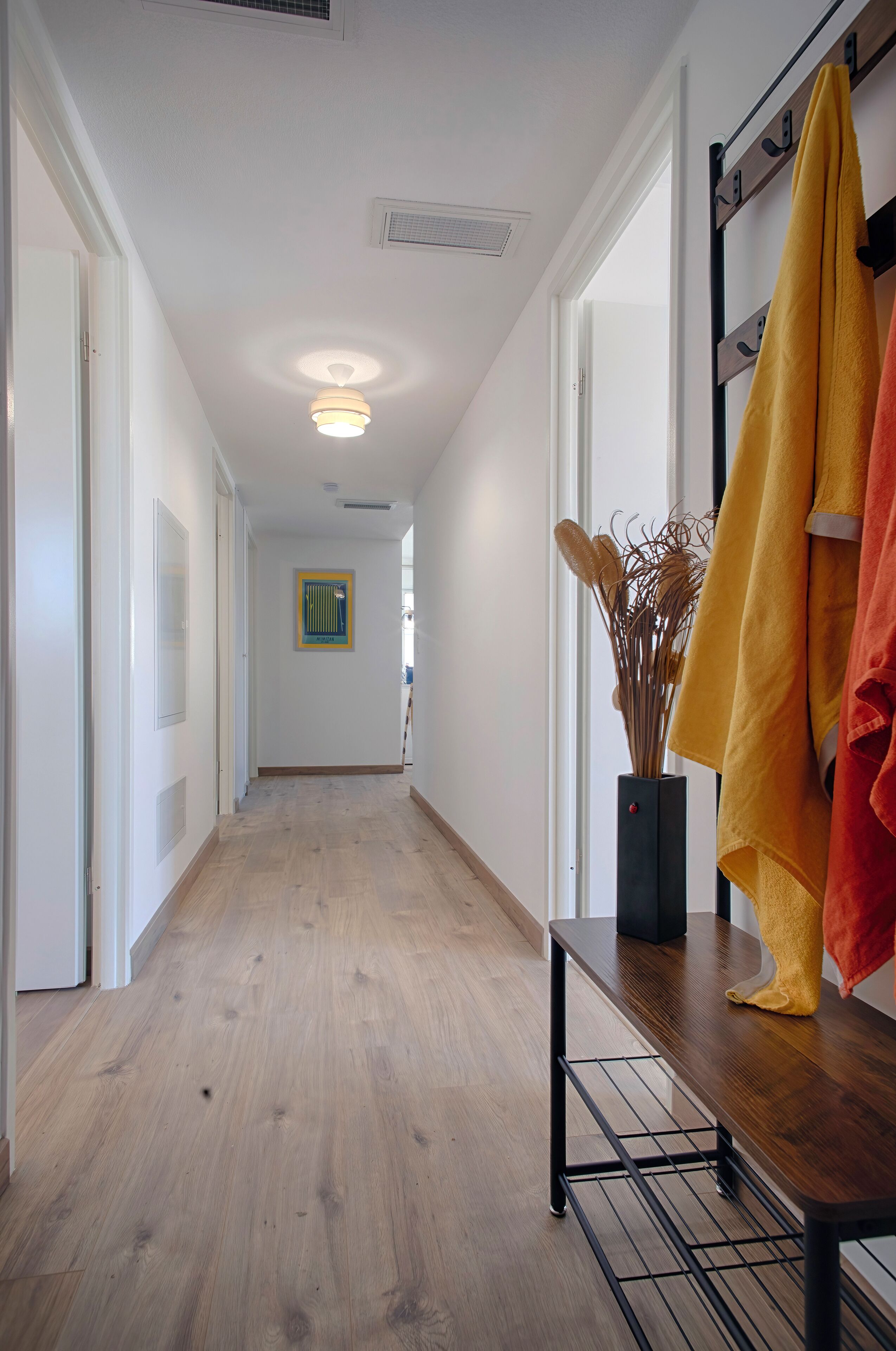 A hallway with wooden flooring, a black metal bench, and a vase with dried plants.