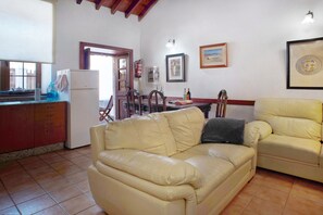 A living room with a beige sofa, a dining table with chairs, a kitchen area with a refrigerator, and a window.