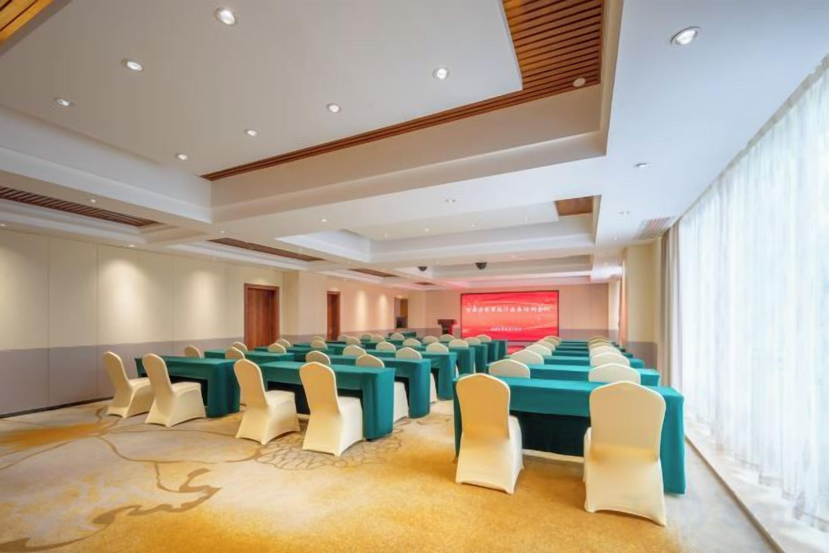 Un salón de conferencias con filas de mesas y sillas, un escenario con un telón rojo y ventanas amplias.
