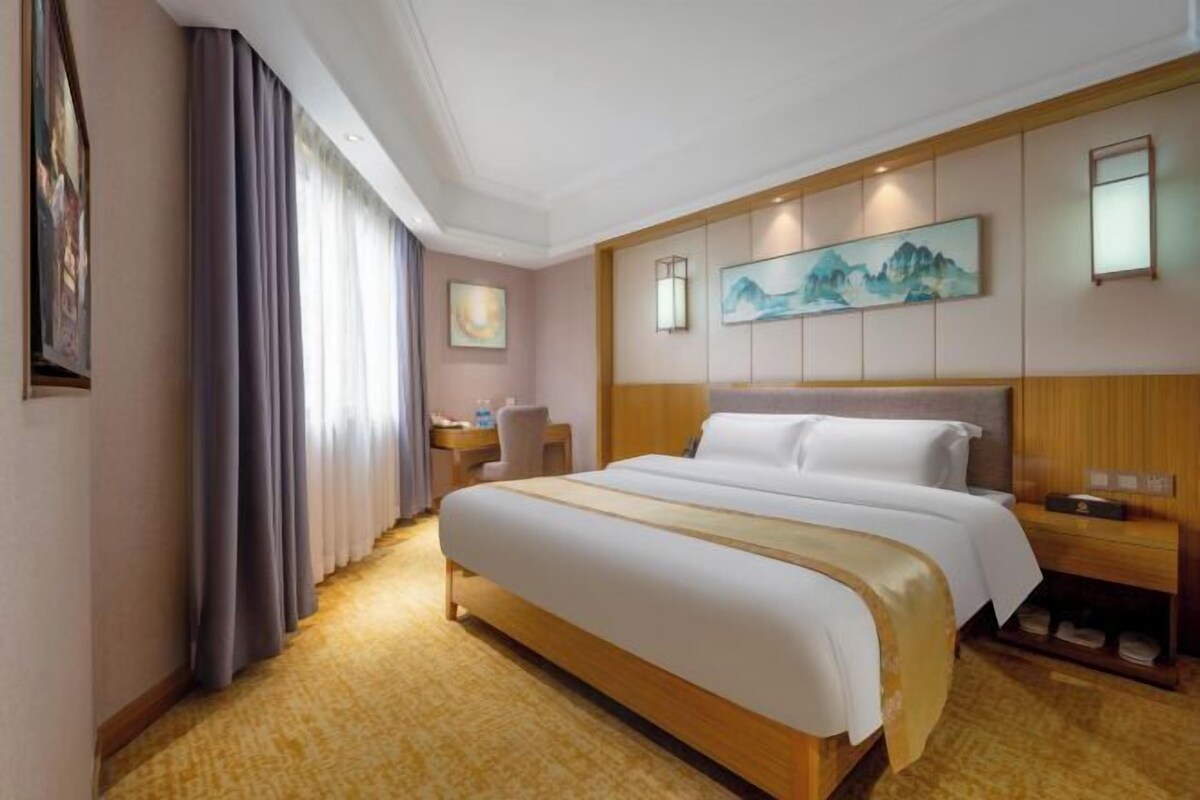 Habitación de hotel con una cama grande, un escritorio y una silla. Hay cuadros en las paredes y una ventana con cortinas.