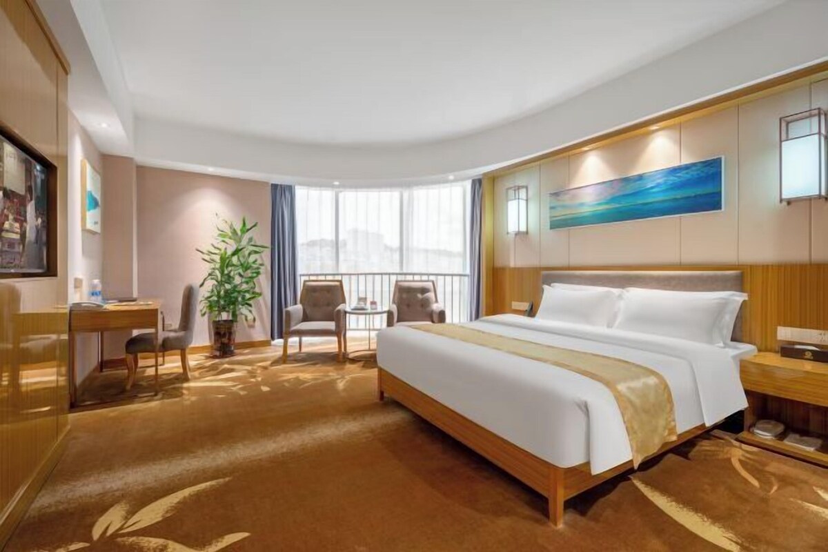 Una habitación de hotel con una cama grande, un escritorio, una silla, una mesita, una planta en maceta y un cuadro en la pared.