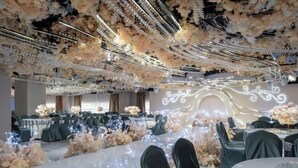 A banquet hall with tables covered in white tablecloths, surrounded by chairs with dark covers. The ceiling is adorned with hanging floral arrangements and lights. - Fusion International Hotel (Yizhang)