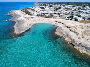 Vista aérea de un pueblo costero con aguas turquesas cristalinas, playas arenosas y un conjunto denso de edificios blancos con detalles azules.