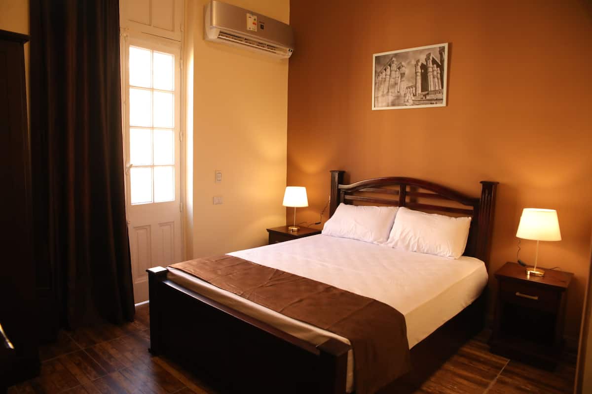 A hotel room with a wooden bed, two bedside tables with lamps, a window with curtains, and a wall-mounted picture.