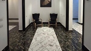 Um corredor com piso de mármore preto e branco, duas poltronas pretas, um candeeiro fixo na parede e uma obra de arte emoldurada.