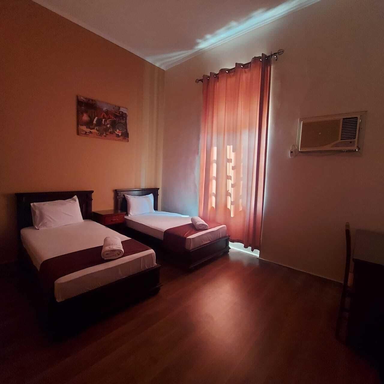 A hotel room with two beds, a painting on the wall, a window with curtains, and an air conditioning unit.