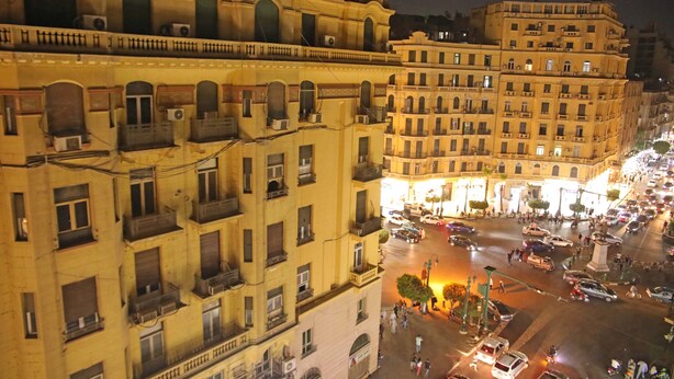Vista noturna de um edifício histórico com vários andares e varandas, cercado por uma rua movimentada com carros e pedestres.