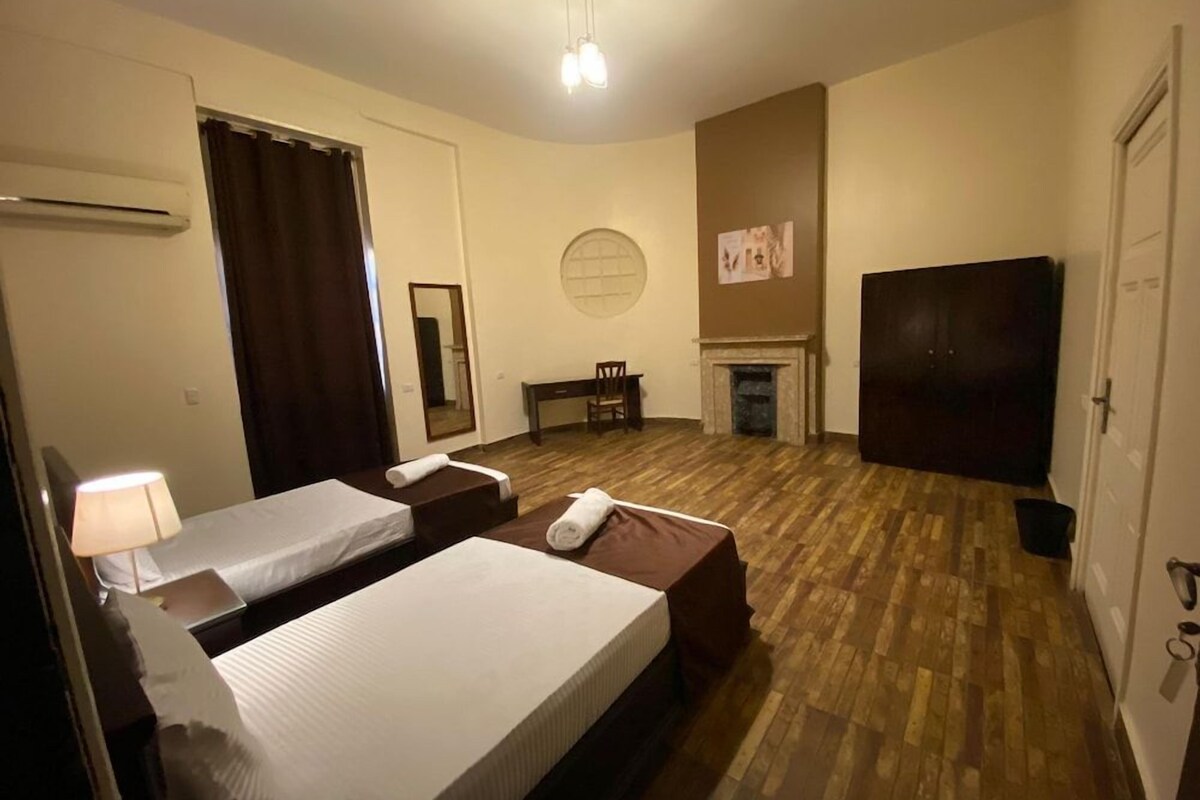 A hotel room with two beds, a desk, a chair, a television, a fireplace, and an air conditioning unit.
