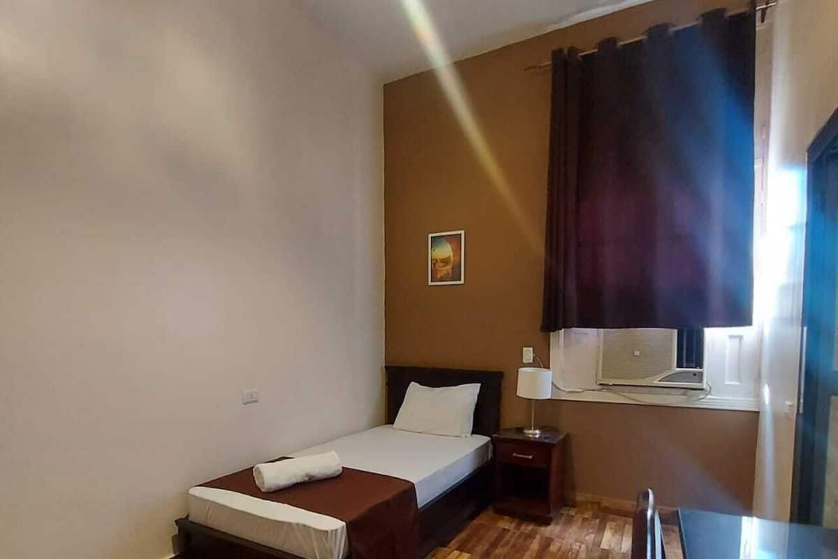 A hotel room with a wooden floor, a bed with a white comforter, a bedside table with a lamp, and a window with curtains.