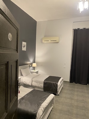 A hotel room with two beds, a wall-mounted air conditioner, a bedside lamp, and a door with a number plate. - Gresham Hotel (Cairo)